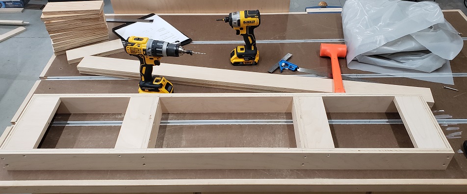 Plywood cabinet base sitting on workbench next to DeWalt drills and orange hammer